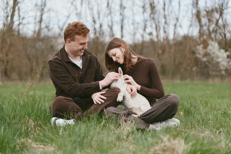A couple sits on the grass in a sunny meadow, smiling as they gently interact with a young goat kid. A vivid lifestyle moment of bonding, warmth, and rural companionship in a natural setting.