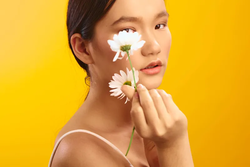 Asian woman holds white flowers against a bright yellow background, a minimalist portrait that radiates calm and elegance, conveying subtle confidence suitable for beauty and fashion imagery.