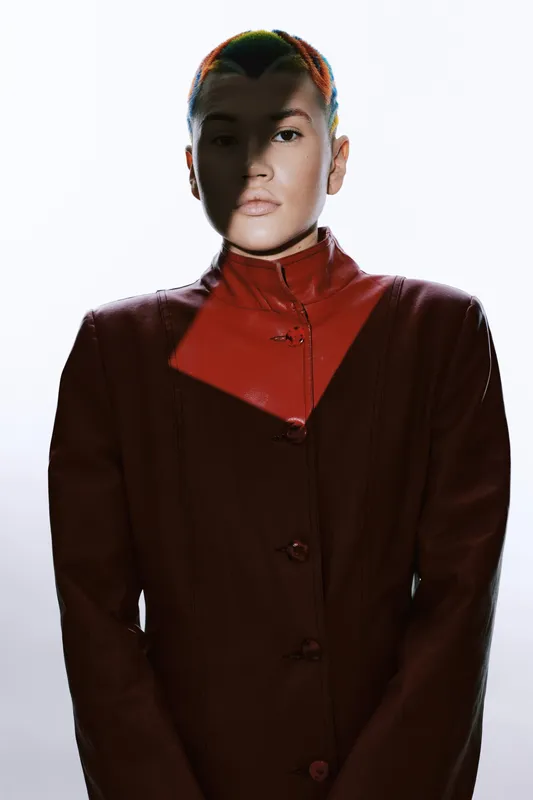 Woman with short hair wearing a dark red high-collar coat, dramatic lighting casting shadows on her face, minimalistic background creating a striking visual impact.