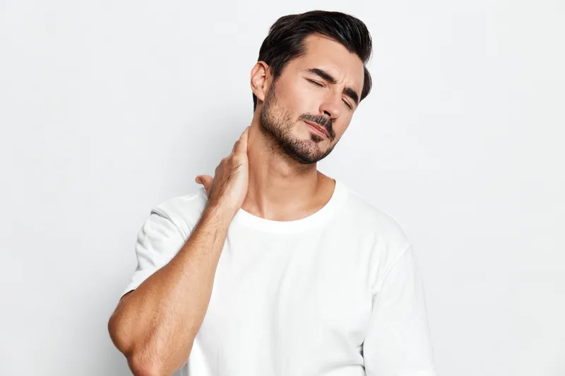 Young man with short dark hair, wearing a white t-shirt, showing a slight discomfort while holding his neck, isolated on plain background. Health concept.