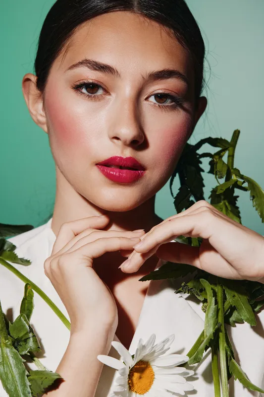 Young woman with styled hair and bright red lipstick in artistic makeup and white clothing poses with green foliage for a beauty and fashion photoshoot with creative concept.