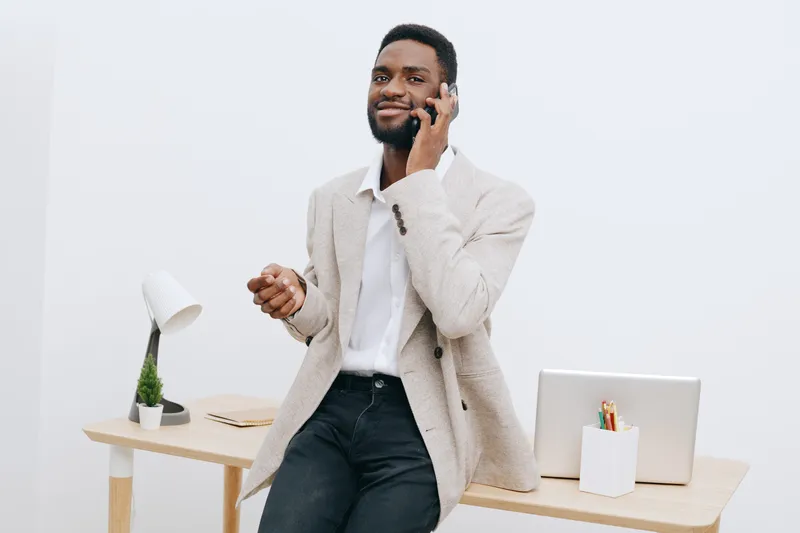 Professional young African American man in smart casual attire talking on mobile phone while standing behind a desk in a modern office setting, looking confident and happy. People lifestyle concept.