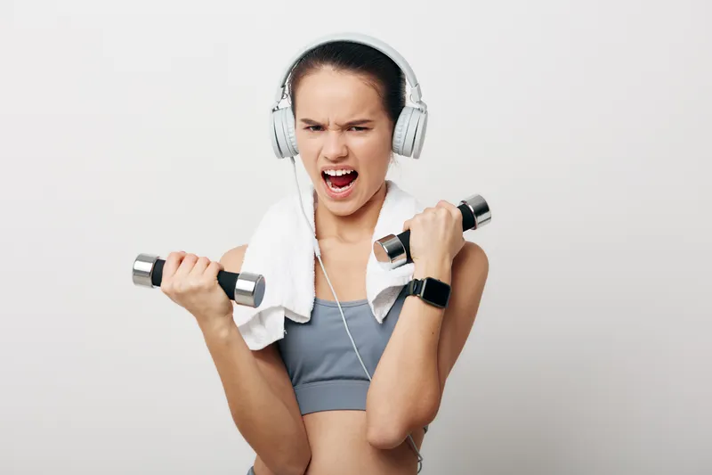 Fitness woman lifting dumbbells with intensity, wearing sports bra and headphones, towel over shoulder in a clean studio, focused energy and powerful workout moment.