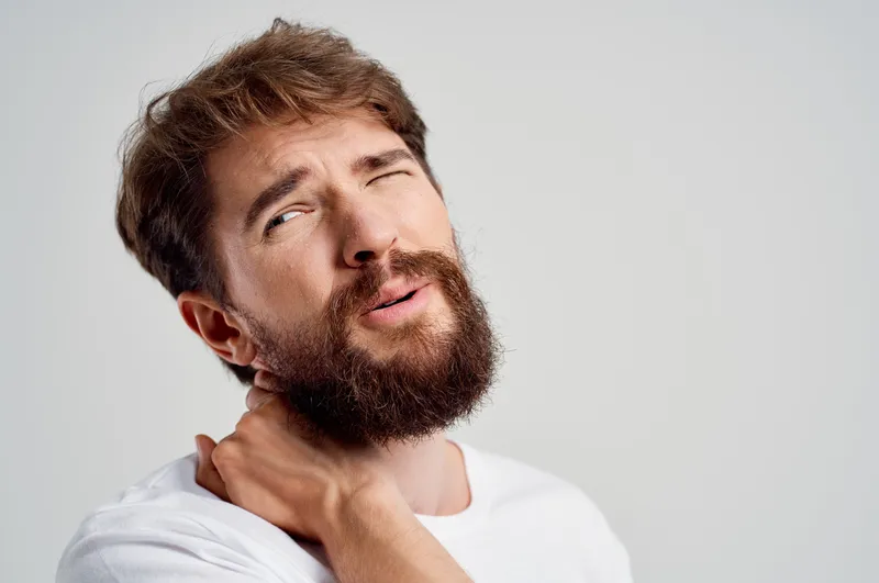 Эмоциональный мужчина испытывает боль в шее, что указывает на проблемы со здоровьем, проходит массажную терапию и лечение в студии