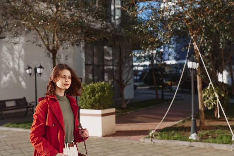 Young woman in a red coat walks thoughtfully along a city sidewalk on an autumn day with trees and street lamps in the background.