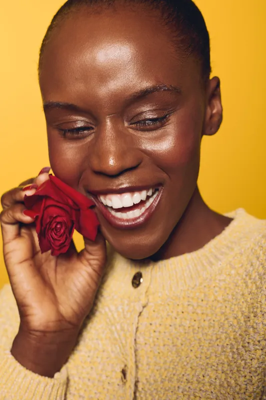 Smiling dark skinned African woman holds red rose near face, wearing yellow sweater. Close-up portrait with yellow background. Concept of happiness, beauty, and natural skincare.