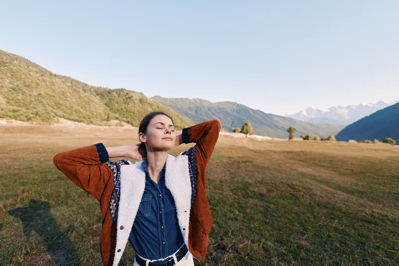 Woman portrait outdoors in a meadow and mountains, relaxed and peaceful with eyes closed enjoying nature and sunlight, casual sweater and jacket conveying freedom and calm.