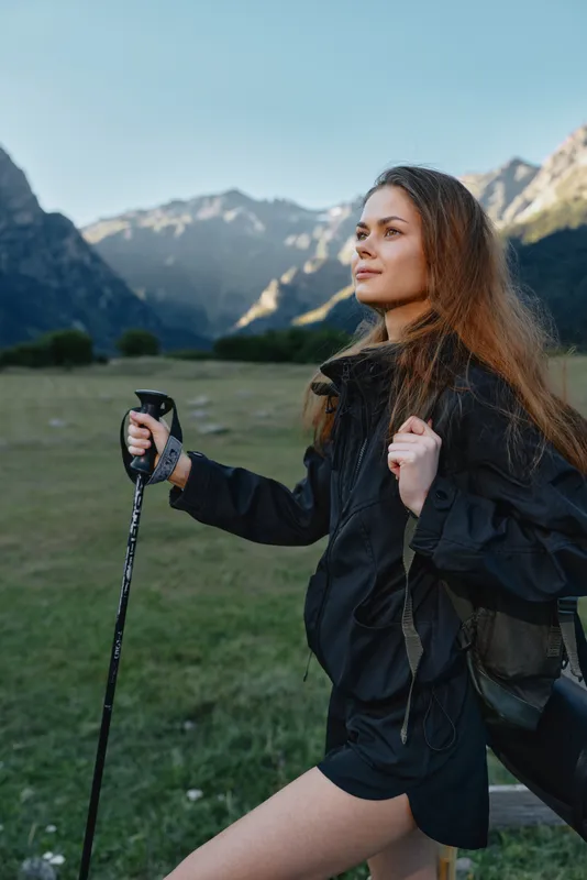 A confident young woman stands in a scenic mountain valley, gripping a trekking pole and looking ahead with calm determination. She wears outdoor gear and embraces adventure in nature.
