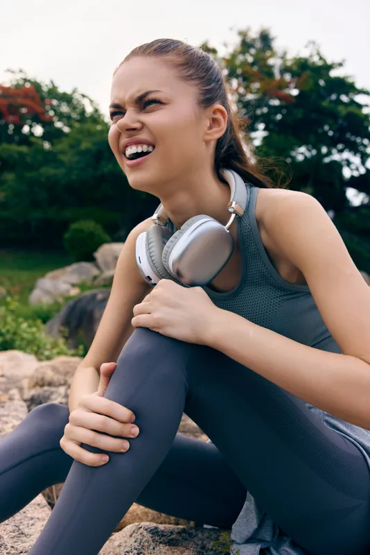 Young athletic female in sporty attire experiencing discomfort while exercising outdoors, wearing headphones, surrounded by nature in a tranquil setting