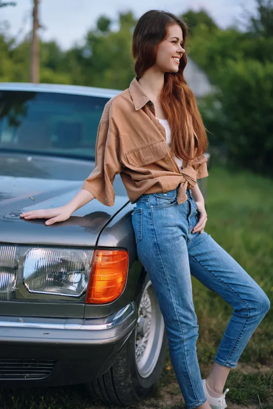Young woman in casual outfit leaning on vintage car, smiling in a green outdoor setting, capturing a sense of relaxation and nostalgia during sunny weather.