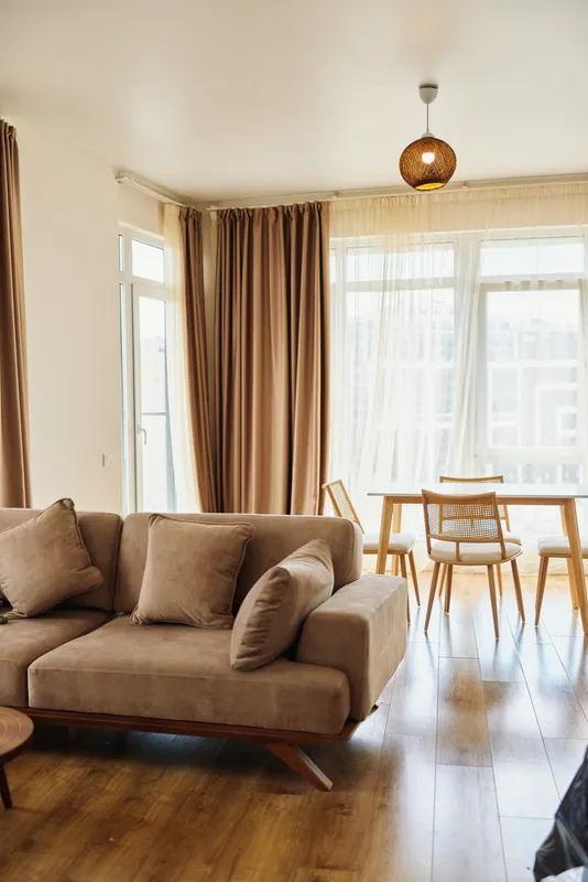 Sofa and cushions in a living area, dining chairs by a big window with light curtains, wooden floor and pendant overhead, bright modern home interior with cozy warm decor.