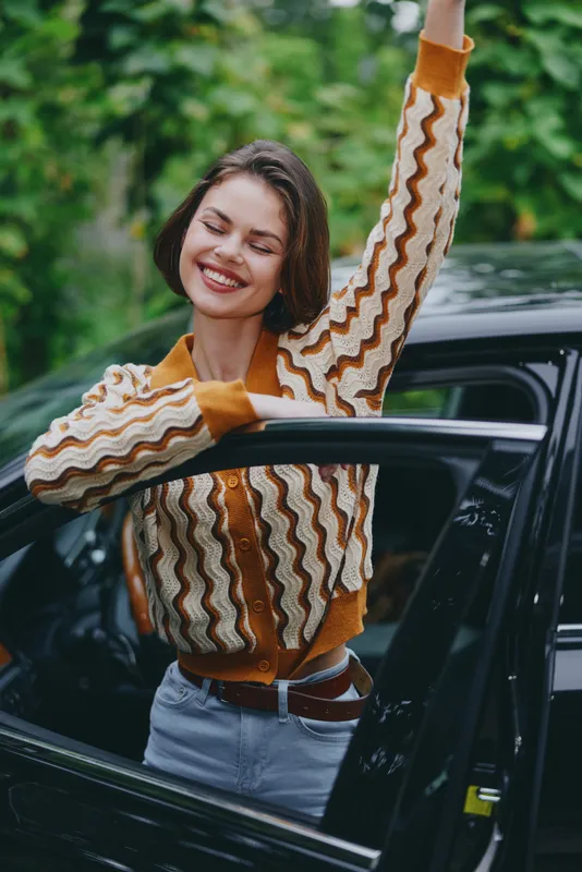 A cheerful woman steps out of a car, smiling with arms raised in a carefree moment against a green park background.