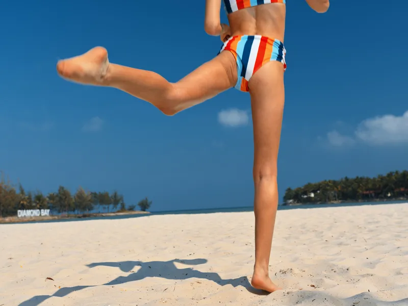 Vibrant scene of a young beachgoer running along sunlit sand in a striped swimsuit, bare legs in motion, captured in a lively action moment under a clear blue sky.