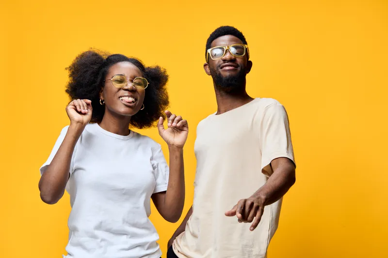 Young African couple dancing joyfully against a bright yellow background, wearing casual clothes and glasses, celebrating happiness and togetherness