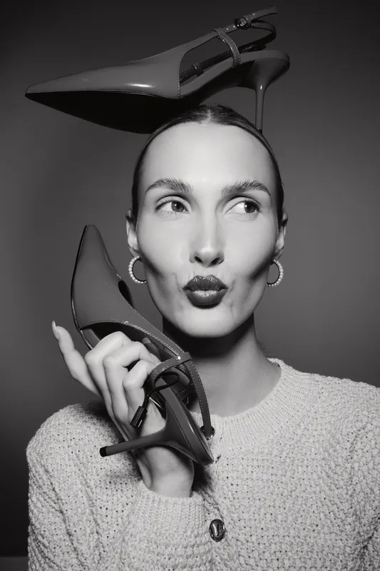 Slim Caucasian woman with light skin balancing high heel shoes on head and hand, wearing sweater, posing with playful expression in studio. Fashion concept.