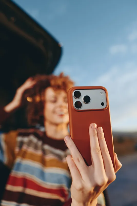 Woman lifestyle smartphone red phone outdoor portrait selfie, curly haired woman holds a mobile in her hand, blurred sky and landscape behind, soft daylight, concept of social connection