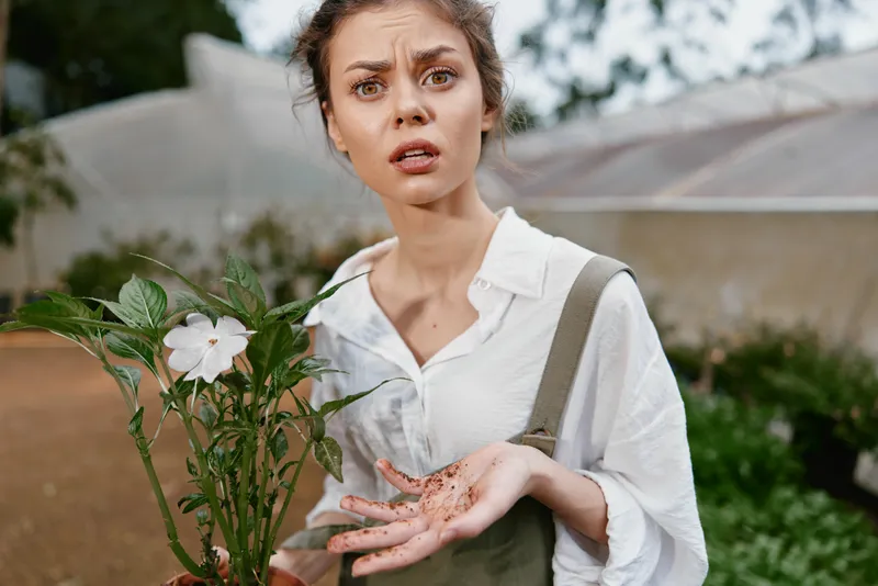 Concerned young woman in gardening attire holds a flower with soil stained hands, reflecting a passion for gardening and the beauty of nature