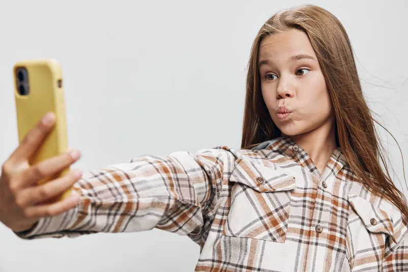 Girl with long hair taking selfie with smartphone in plaid shirt against white background. Portrait, expression and casual style for social media content creation.