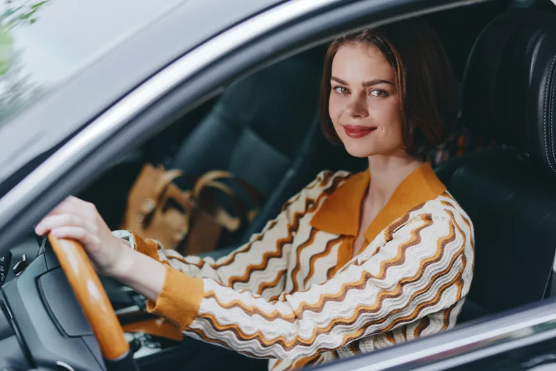 A confident woman sits behind the wheel, smiling, wearing a retro striped shirt and warm tones.