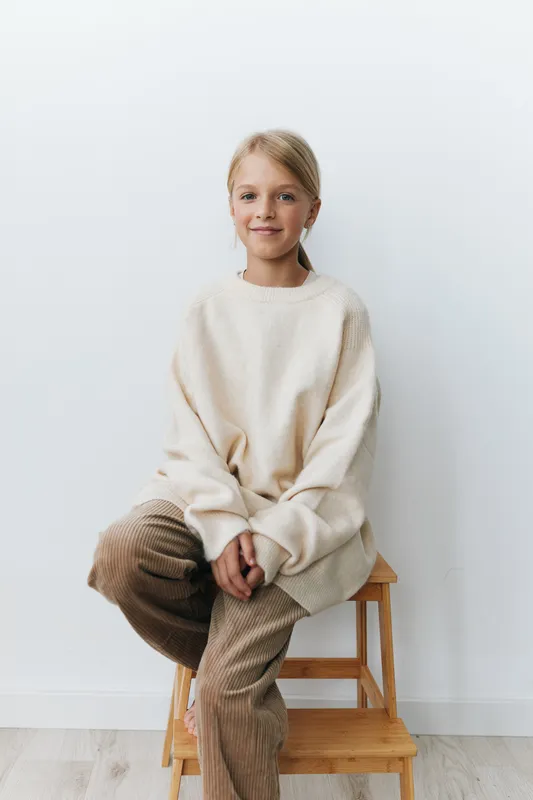 A young girl sits on a wooden stool wearing an oversized cream sweater and corduroy pants, posed calmly against a clean white backdrop.