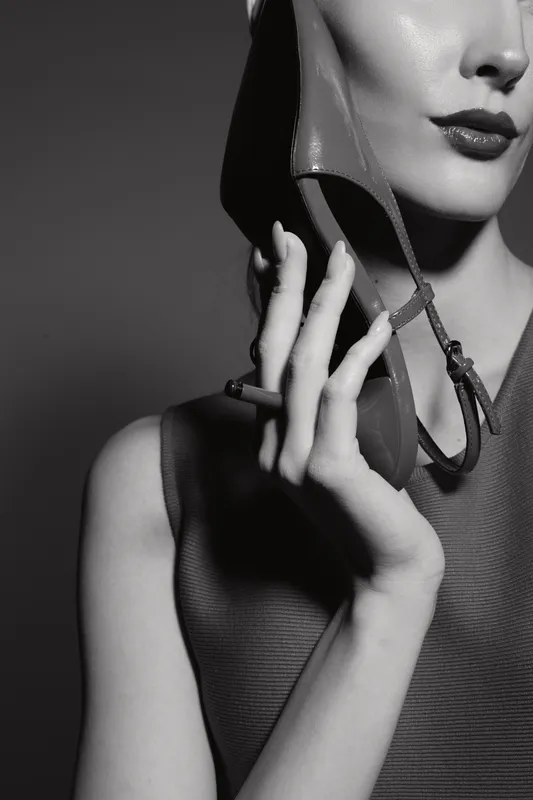 Close-up of a pale white woman with light skin holding a high heel shoe near her face, lips painted dark, cigarette in hand, black and white fashion concept with retro style.