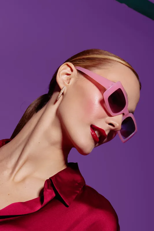 Stylish woman with pink sunglasses and red lipstick poses against purple background. Fashion portrait with glossy makeup and sleek hairstyle. Concept of beauty, style, and modern elegance.