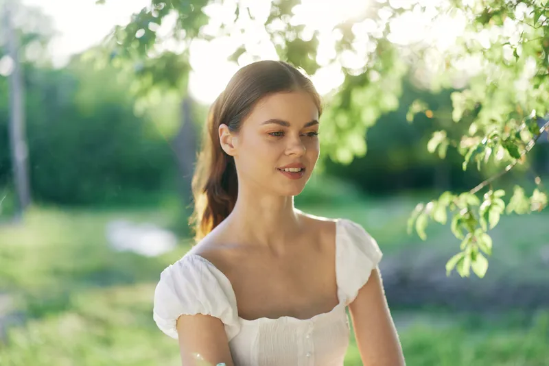 Young woman in a white dress smiling gently in a sunny green park, embodying tranquility and natural beauty, perfect for lifestyle and wellness themes