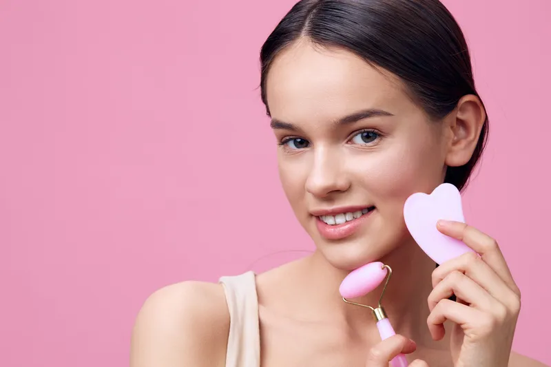 Young woman with a facial roller and heart shaped beauty tool on a pink background, promoting skincare and self care routines for healthier skin