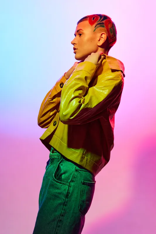 woman with colorful shaved hair posing confidently in a yellow jacket and blue jeans against vibrant pink and purple gradient background, modern fashion style