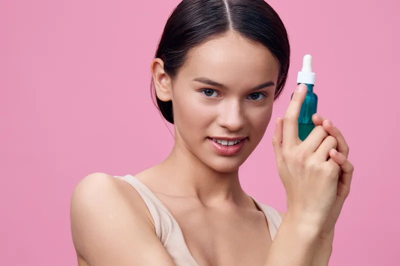 Young woman holding a serum bottle with a confident smile, showcasing beauty and skincare products against a vibrant pink background Perfect for healthy skin concepts