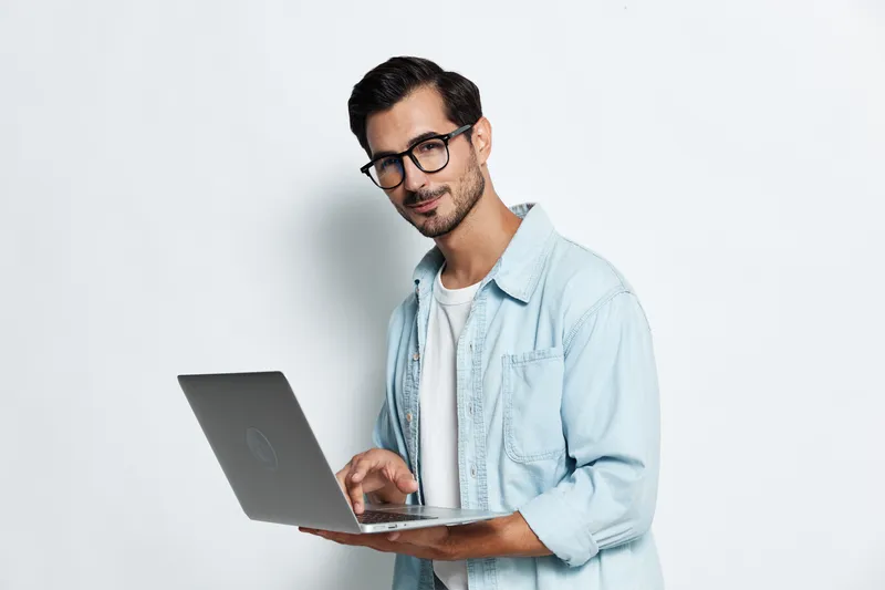 Smiling man wearing glasses and a light denim jacket, holding a laptop while posing against a plain solid background. Professional lifestyle concept.