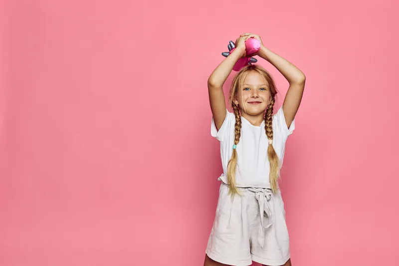 Happy girl in white t-shirt and shorts holds pink dumbbells above her head on a pink background. Concept of children fitness and active lifestyle.