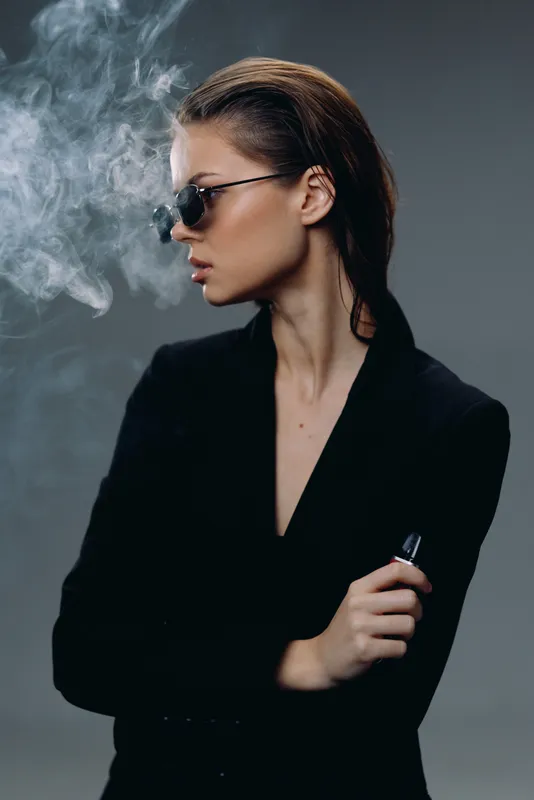 Stylish woman vaping with sunglasses blowing smoke in a dark studio setting.