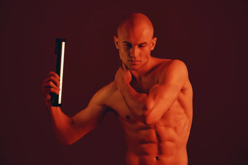 A muscular man posing with a light source, creating a dramatic red ambiance, showcasing strength and allure in an artistic black and white setting.
