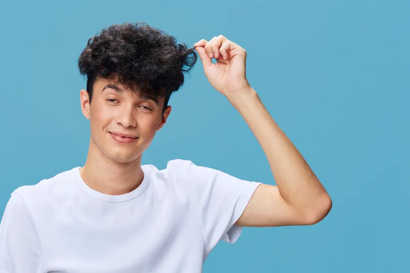 young man in white t-shirt playfully pulling curly black hair against blue background, smiling with eyes half-closed, casual and fun portrait, light pastel colors
