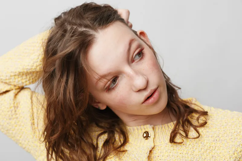 Young woman with curly brown hair wearing yellow sweater looks sideways with thoughtful expression on light background. Concept of contemplation, casual style, and natural beauty.