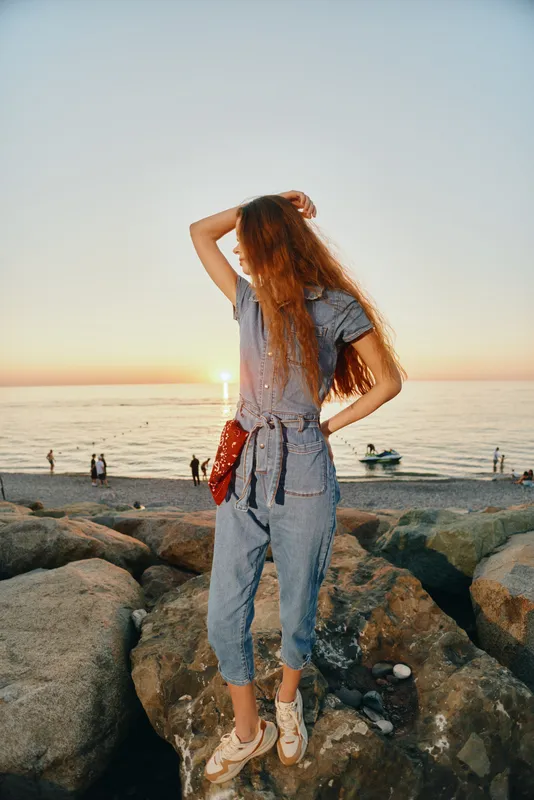 Lifestyle woman in modern boho-western denim street style posing on rocks at sunset, film color with relaxed beach atmosphere and casual fashion vibe.