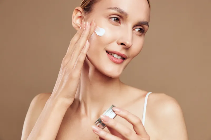Young woman with clear skin applies moisturizing cream on her face while smiling gently against a neutral background in a skincare routine moment.