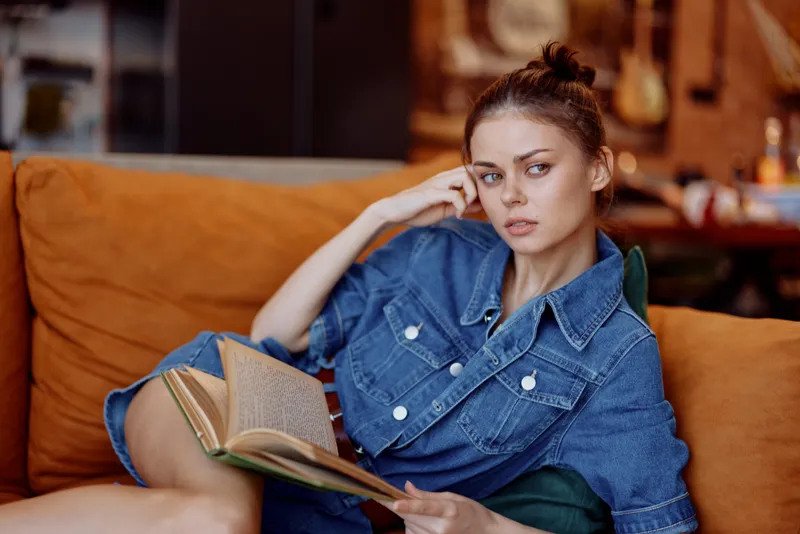Молодая женщина в джинсовом платье сидит на диване, увлечённо читает книгу и уверенно смотрит в камеру