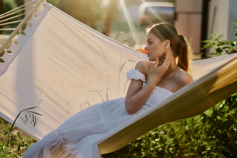 Young woman in a white summer dress relaxing in a hammock during golden hour, showcasing tranquility and serenity in a natural garden setting