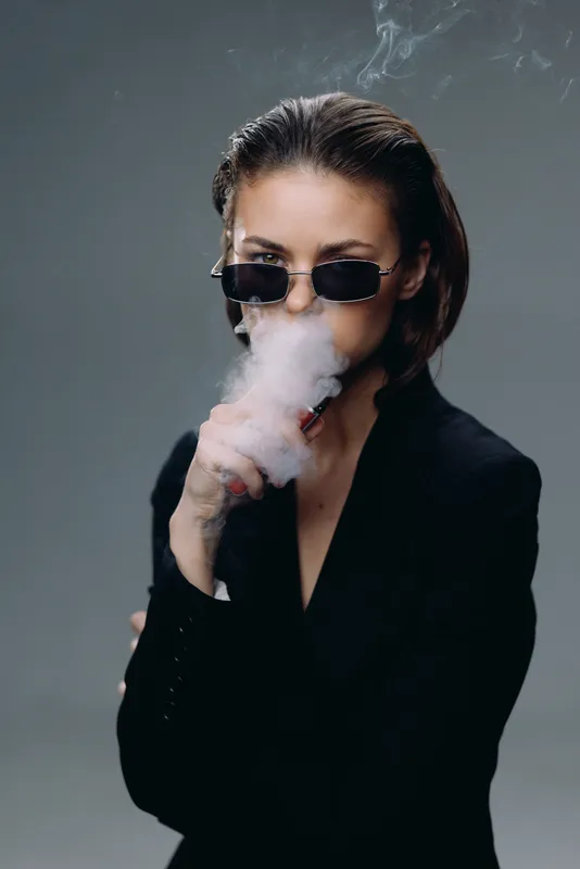 Woman vaping with sunglasses in dark outfit, smoke around face, moody indoor portrait with gray background