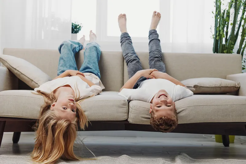 Two happy children lying upside down on a modern sofa in a bright living room, smiling and having fun together. Concept of childhood joy and playful moments.