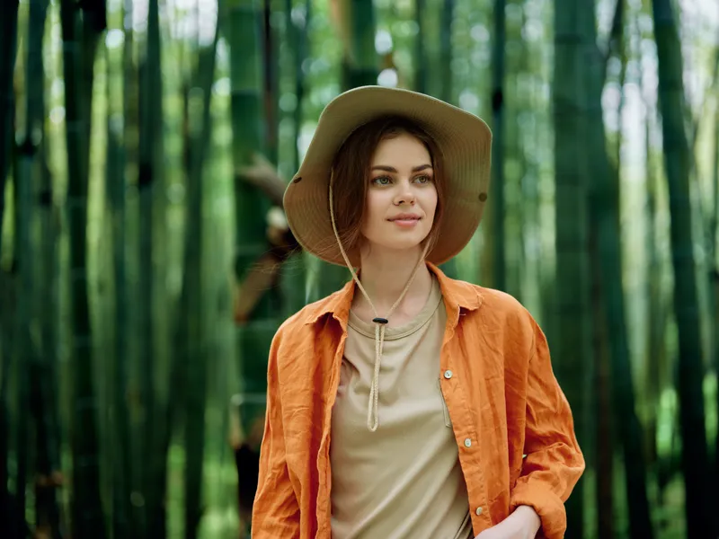Woman portrait in bamboo forest wearing wide brim hat and orange jacket, travel lifestyle and environmental portrait with serene expression, nature walk, green stalks and soft natural light,