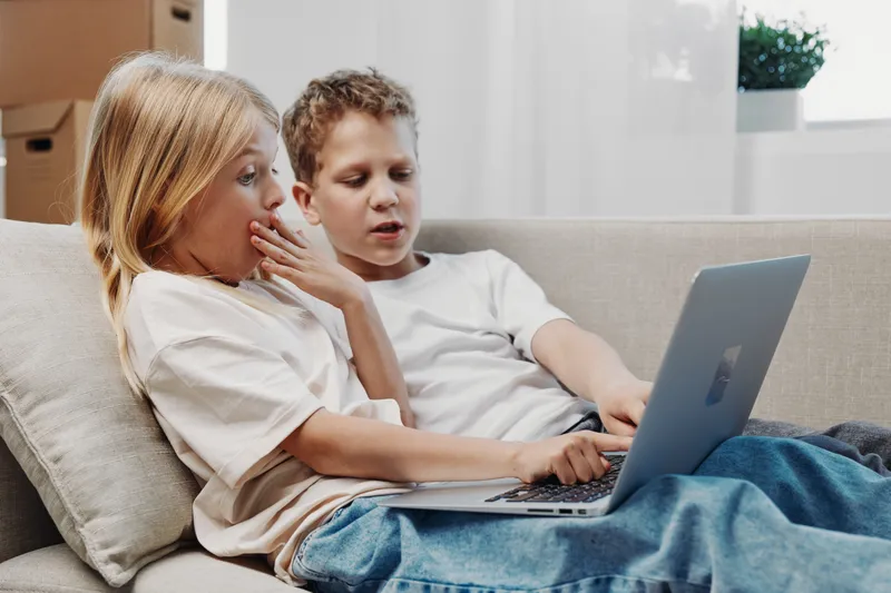 Two children in casual clothes sitting on sofa watching laptop screen with curious and surprised expressions in bright living room.