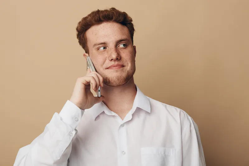 Young man in white shirt talking on smartphone, looking away with slight smile. Neutral beige background. Concept of communication, technology, and casual lifestyle.