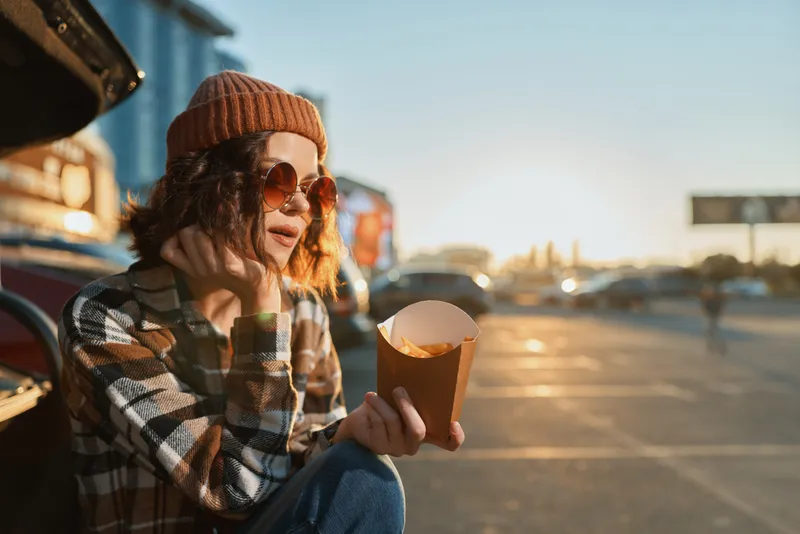 Woman eating fries by a car at sunset, wearing beanie and sunglasses in a casual street scene, candid lifestyle and authenticity with golden hour glow and emotional storytelling.