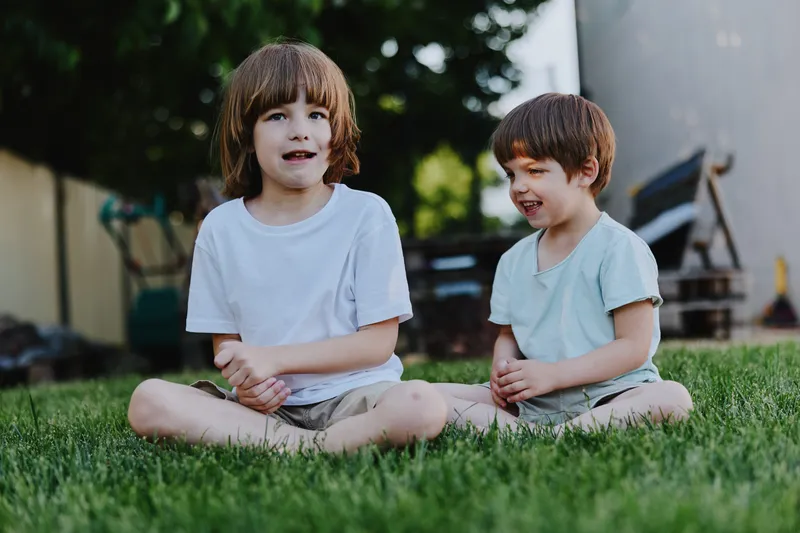 Two cheerful boys sitting on green grass, enjoying a sunny day outdoors, dressed casually in comfortable summer clothing, radiating joy and playfulness.