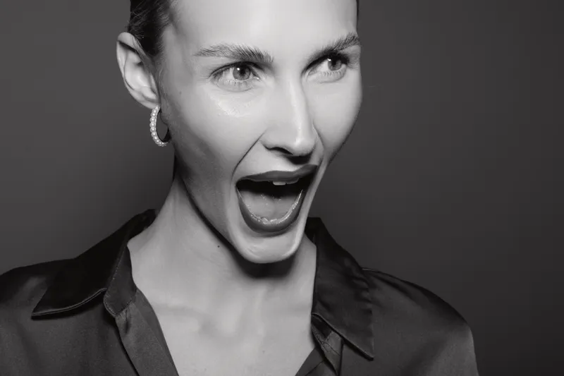 Young Caucasian woman with light skin and short hair looks surprised and excited, wearing hoop earrings and a silk shirt in black and white studio portrait.