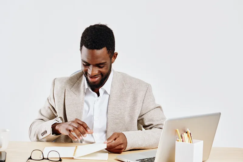 Confident smiling man in beige blazer and white shirt working at desk with open notebook, glasses, and laptop on plain white background studio portrait. Business success concept