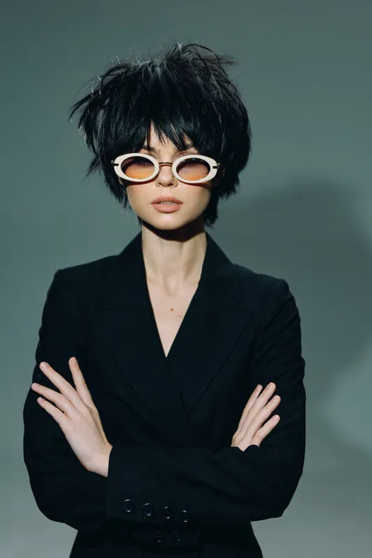Stylish woman with short black hair and sunglasses wearing a black outfit against a gray background.